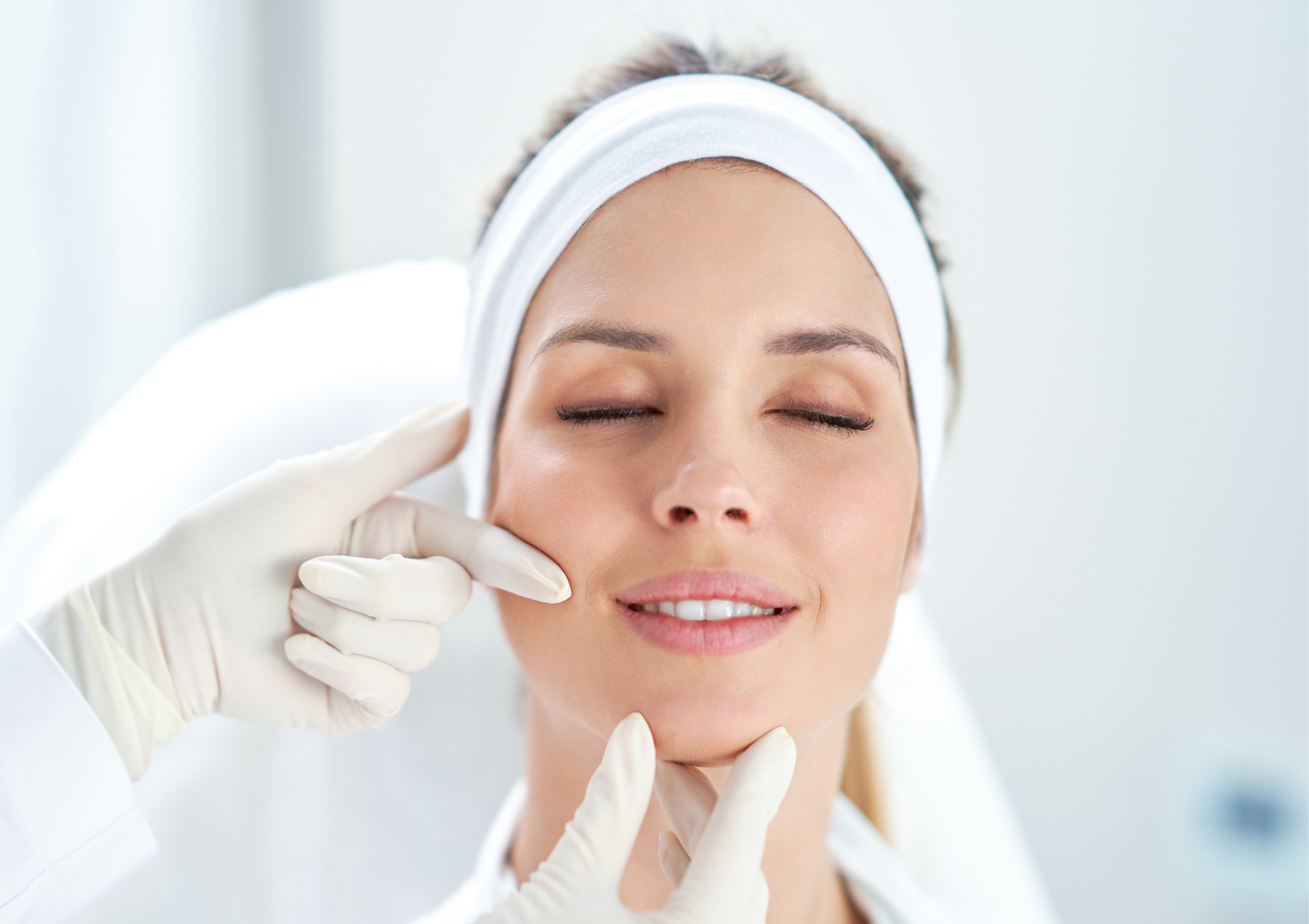 Close-up of a woman with a white headband having a facial treatment; gloved hands gently lift her chin and cheeks with a soft touch.