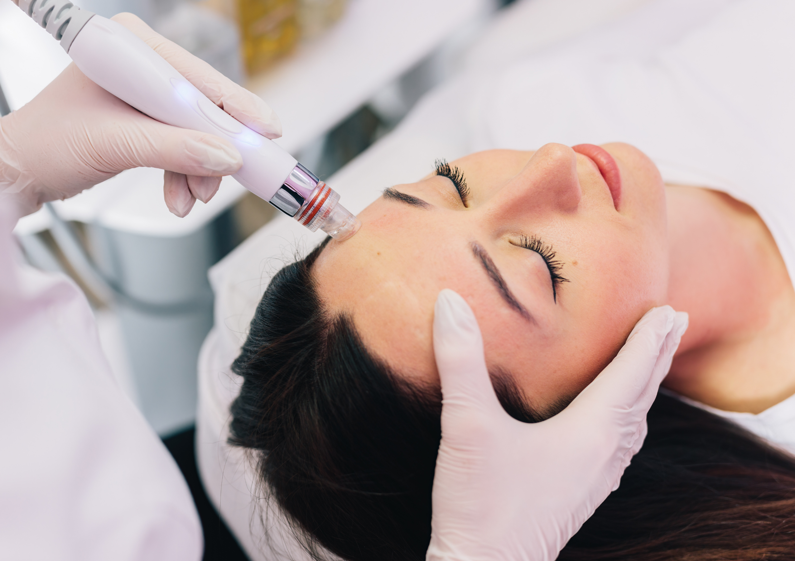 Clinician in gloves uses a handheld cosmetic laser on a woman's cheek during a facial treatment.